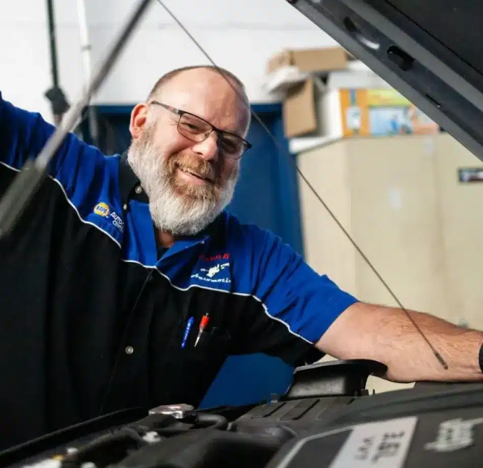 A smiling mechanic with a white beard and glasses works under the hood of a car in a garage, wearing a blue and black uniform with pens in his pocket.