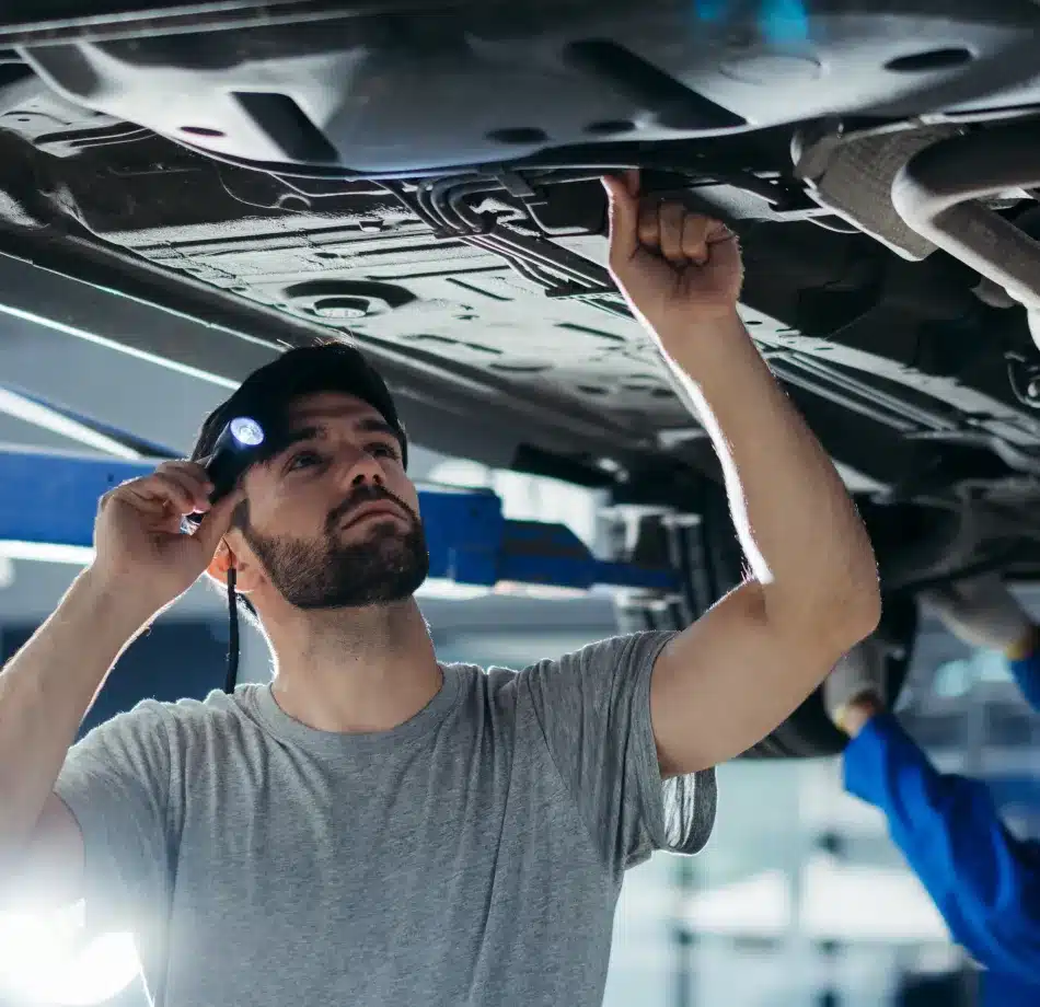 Cain and Sons’ Automotive technician wearing a grey t-shirt and headlamp inspecting a vehicle’s underside for ADAS calibration in Glen Rock, PA, with another mechanic working nearby.