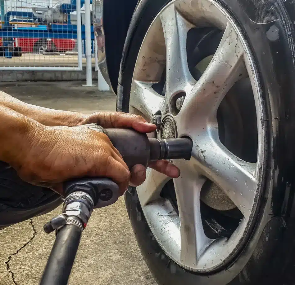 Close-up of hands using a power tool to tighten lug nuts on a car wheel. The setting appears to be a garage, conveying focus and precision.