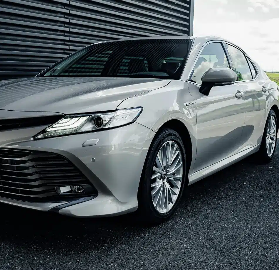 Silver sedan parked on asphalt with a modern, sleek design. The car's LED headlights are on, with a metallic building wall and a cloudy sky in the background.